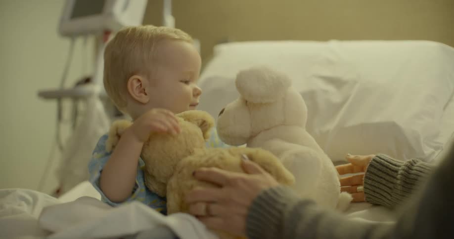 Woman gives toddler child sitting on the hospital bed a teddy bear in the hospital