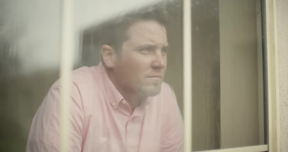 A man looks out a window and bows his head down