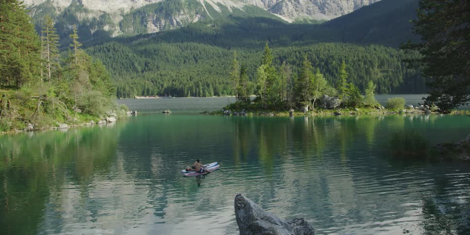 A person sits in a canoe or boat on a calm lake surrounded by trees and mountain