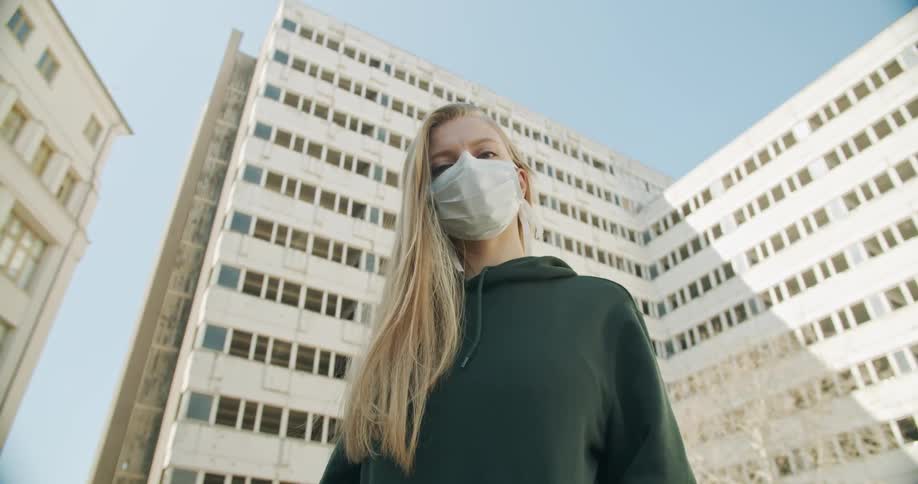 A woman stand outside of a large building wearing a medical face mask