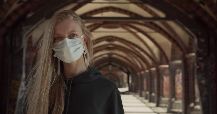 Blonde woman wearing medical mask stands in front of arched roof over walkway