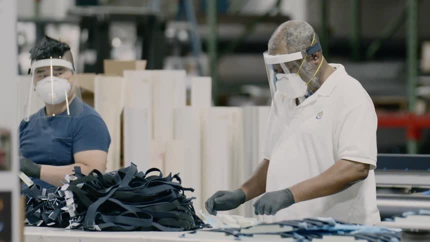 Workers construct product on table while wearing masks and face shields