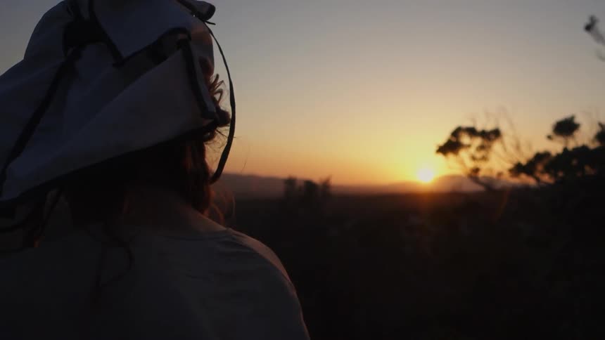 A bearded man wearing a hat, looking around and stepping to the side as the sun sets in the mountains