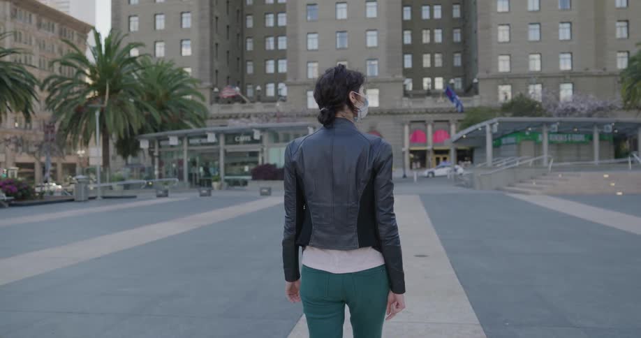 Woman walks toward building with shops on the ground floor wearing medical mask