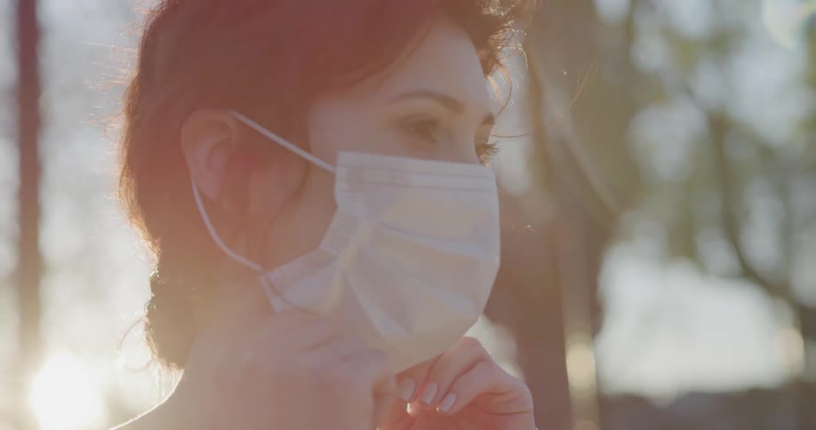 A woman standing outside on a bright sunny day puts on a mask