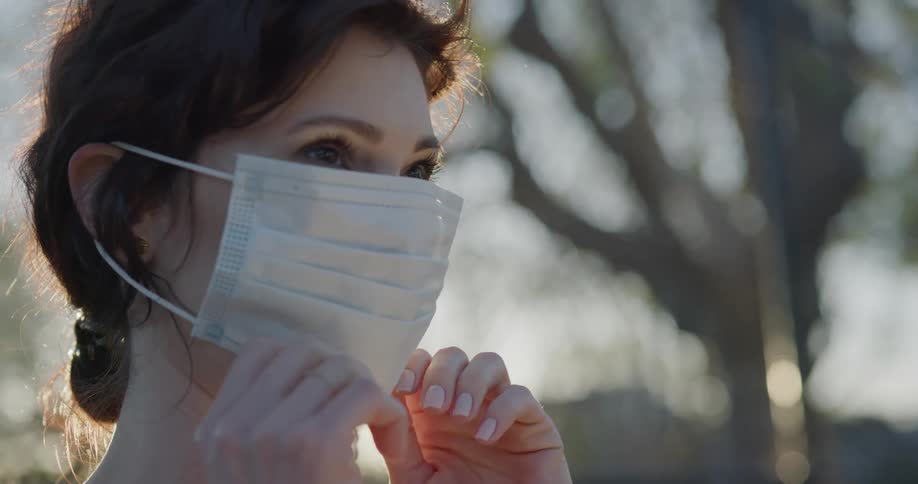 A woman standing outdoors puts on a surgical mask