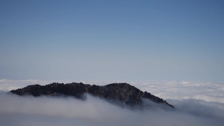 The top of a mountain peeks through many fluffy white clouds with a blue sky