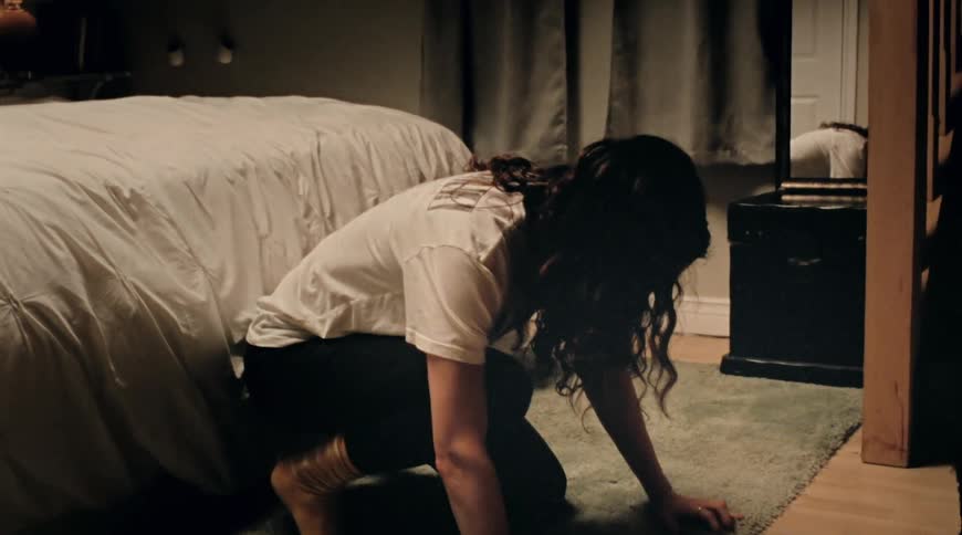A woman on one knee leaning forward on her hands in her bedroom turns her head to look at her reflection