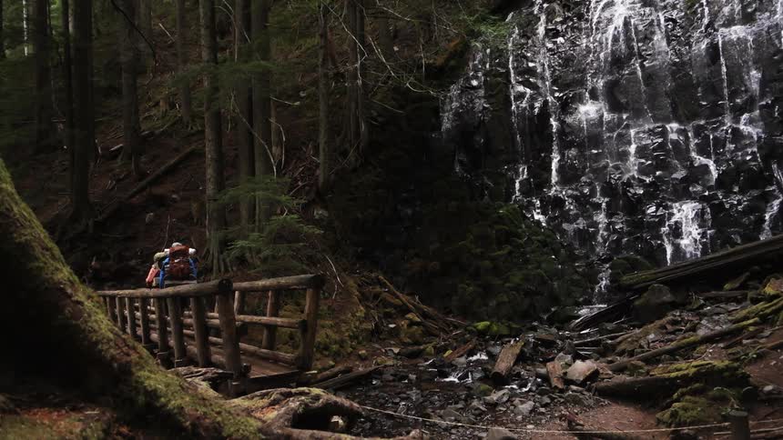 Hikers crossing a wooden bridge as a waterfall trickles down the face of a cliff off to the side in the woods