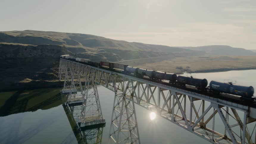 Cargo train crossing an enormous steel bridge spanning a calm river reflecting the sun