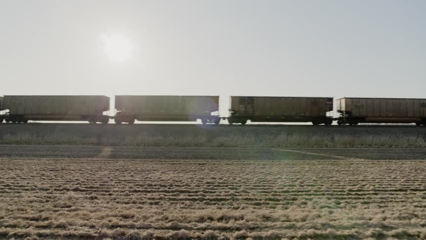 Backlit cargo train hopper cars traveling on a raised track along a plowed farm field