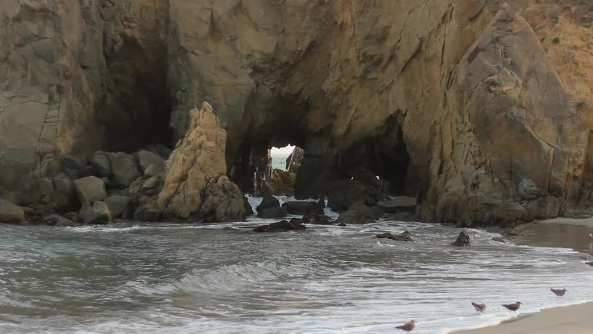 Birds along the shoreline as waves roll in around, and through, a large rock formation