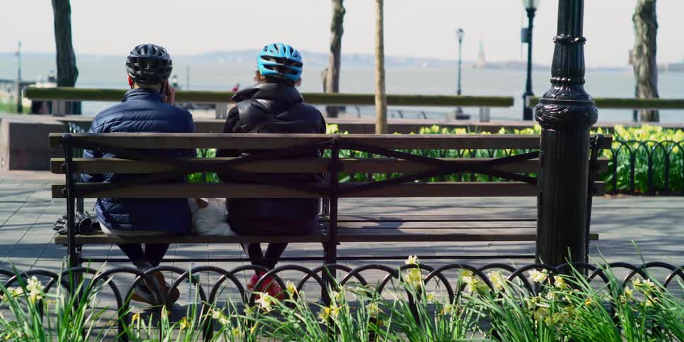 Two people sit on a park bench wearing helmets