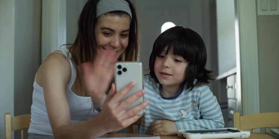 A mom and son at the kitchen table wave and smile during a video call on a smart phone