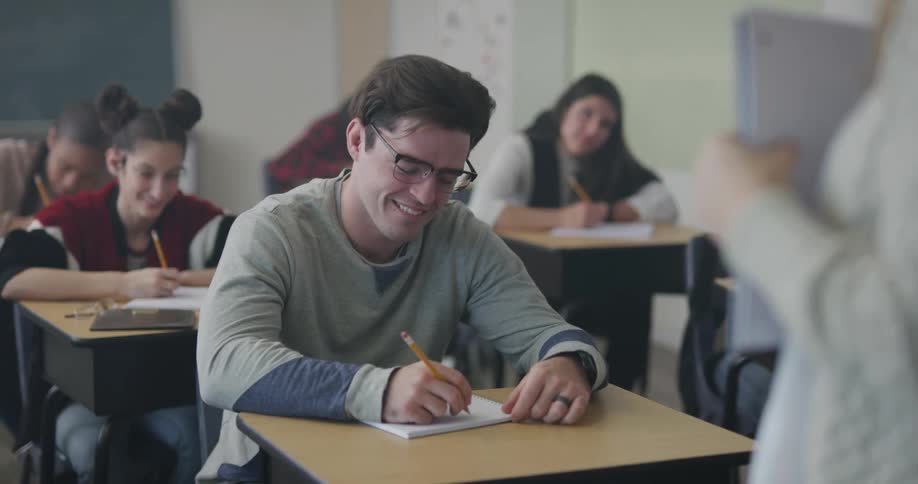 A teacher standing in front of the class talking while students smile and write