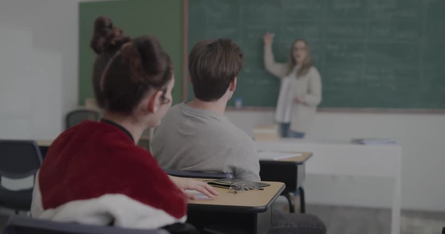 Students in a classroom while the teacher stands in front of the blackboard teaching