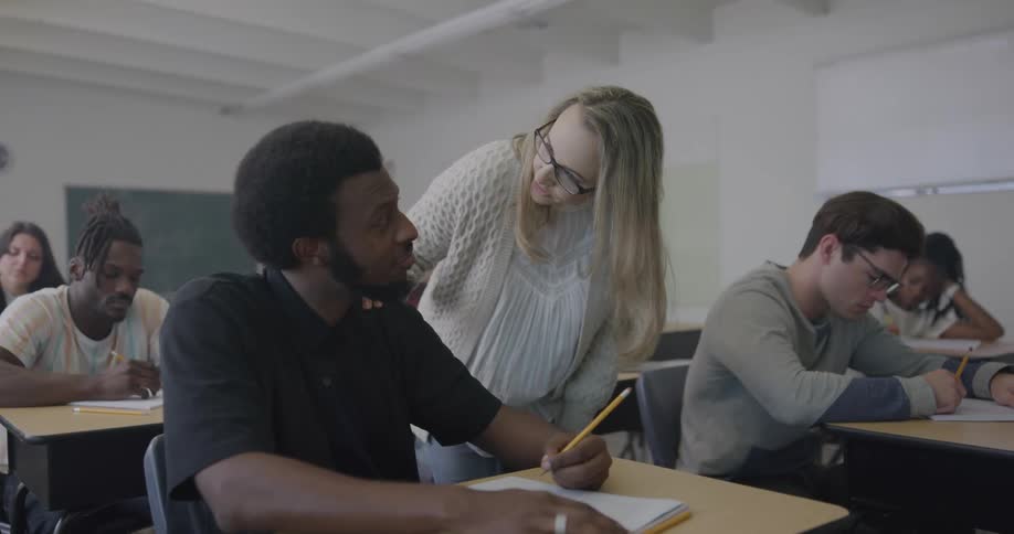 Students in a classroom sitting and writing with a teacher talking to them