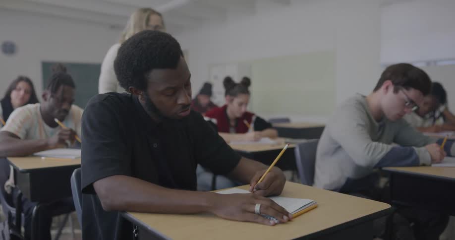 Students in a class sit at their desks all working and writing as the teacher walks around checking in with them