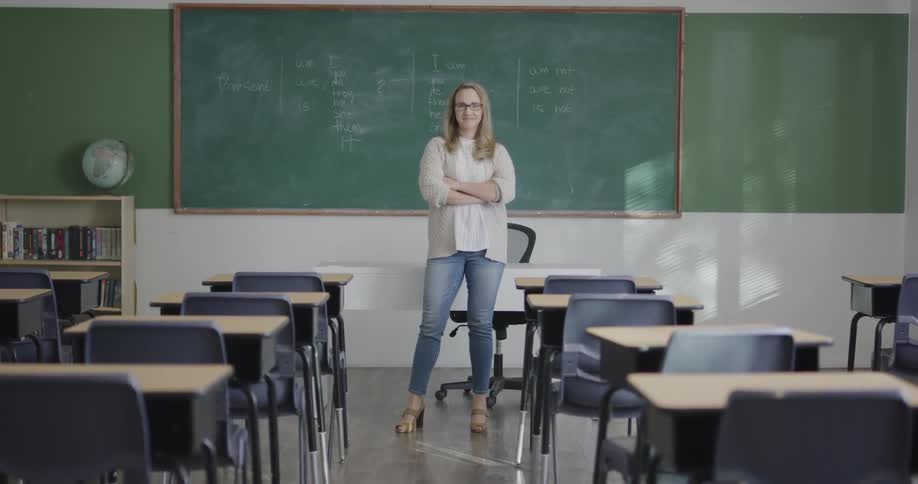 A teacher walks from behind her desk and stands at the front of a classroom and crosses her arms