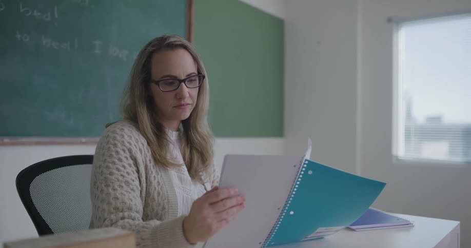 A teacher sits at the front of a classroom and looks in a notebook