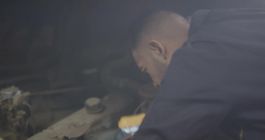 A mechanic holds a work light as he is bent over under the hood of car