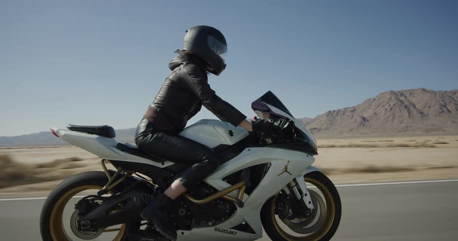A woman riding a motorcycle on a desert road pulls up next to another vehicle turns her head to look then speeds ahead