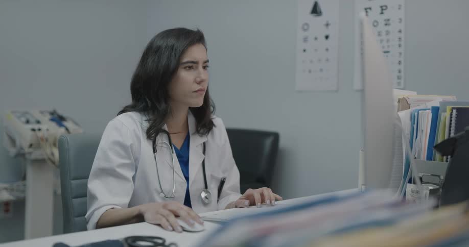 Doctor sits at desk entering data into computer from paper