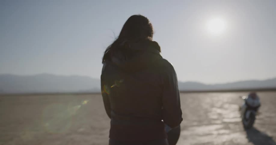 A woman in leather putting on her helmet as she walks through a desert towards a motorcycle