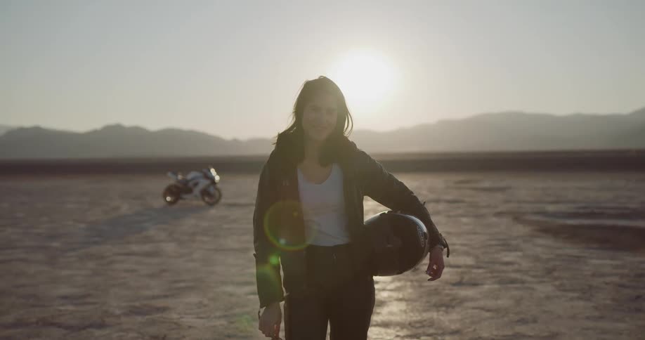 A woman stands in a desert area holding a motorcycle helmet