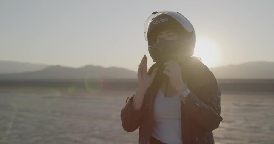 A woman dismounts her motorcycle and takes off her helmet in the desert at dusk