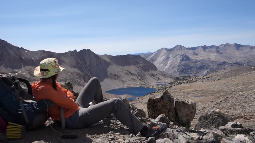 A woman sitting on rocky ground, leaning back against a backpack and looking out over mountains, a valley and a body of water on a windy day