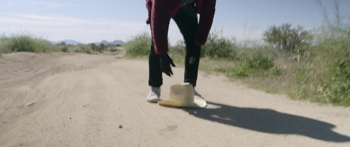 A man wearing a black full head and face cover staggers backwards on a dirt road in the desert dropping his hat and picking it up