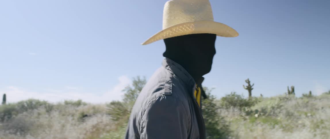 Man wearing black face mask and cowboy hat walks through desert
