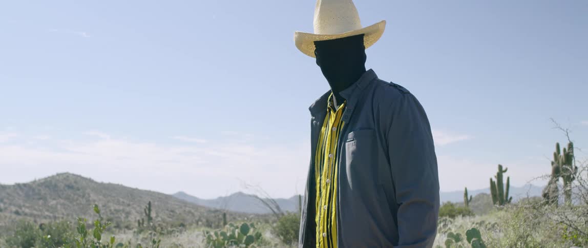 Man wearing a black face mask and cowboy hat stands on desert