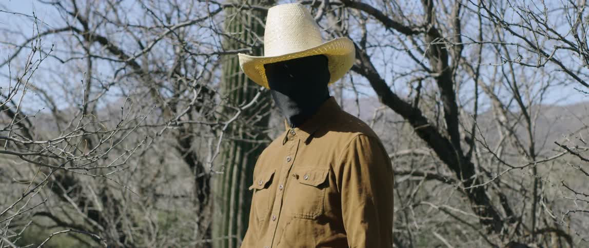 A man wearing a black full face and head covering under his cowboy hat stands near a cactus and trees