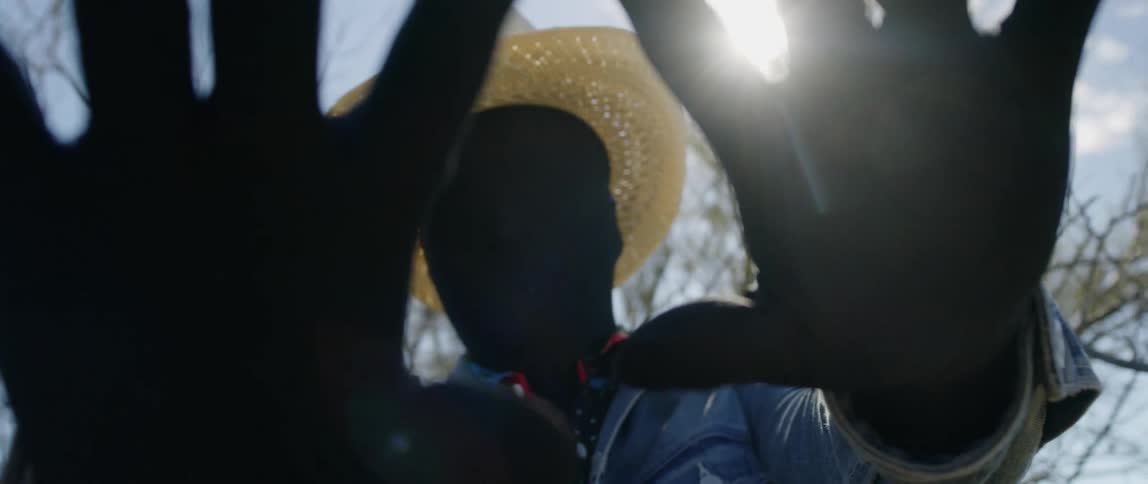 A man wearing a black full head and face cover and black gloves holds his hands out and palm up on a sunny day