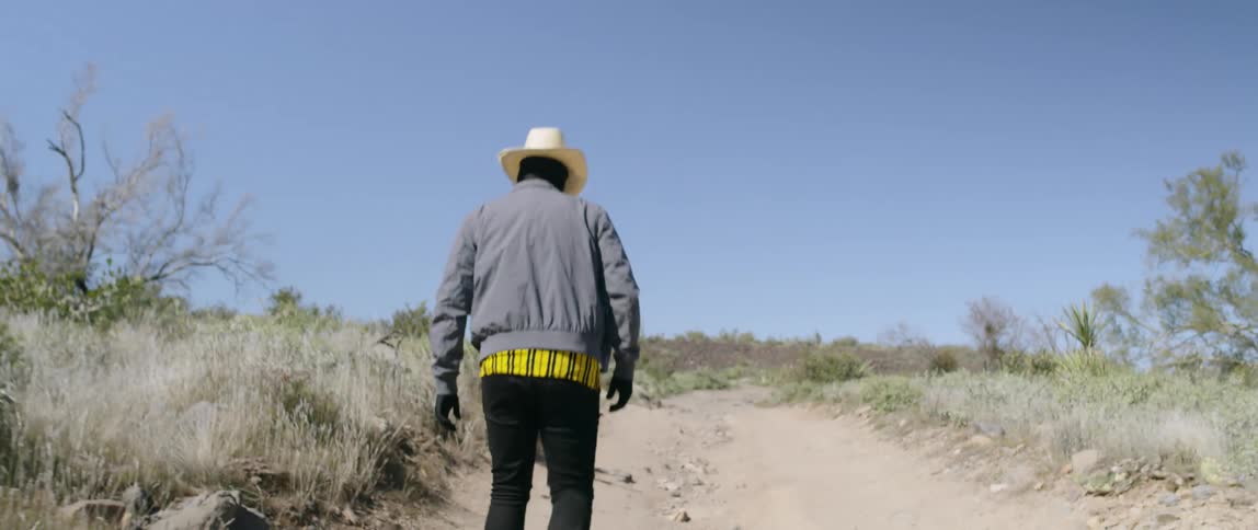 Man wearing black face mask and cowboy hat walks on a path on the desert
