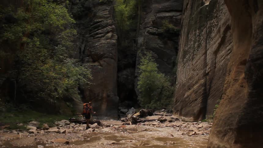 A person hiking alongside a shallow, rock-strew stream flowing between steep cliffs in a wooded area