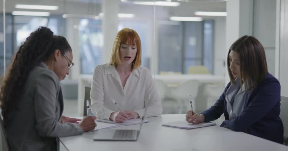 Three women sit at a table in a conference room and talk and take notes