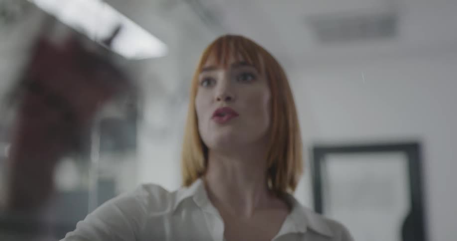 A woman stands in an office writing on a clear dry erase board talking and brainstorming with two other women