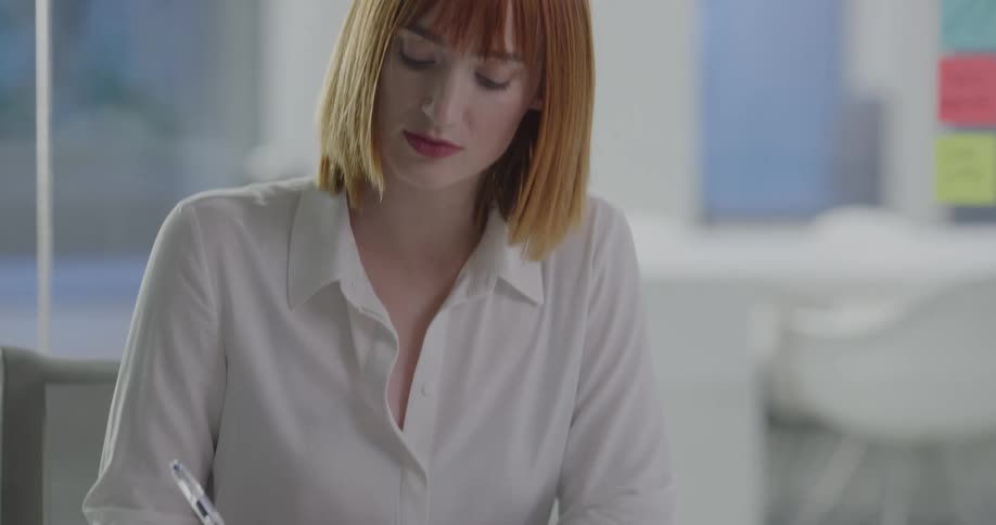 Red haired woman sits at table writing on notepad in office
