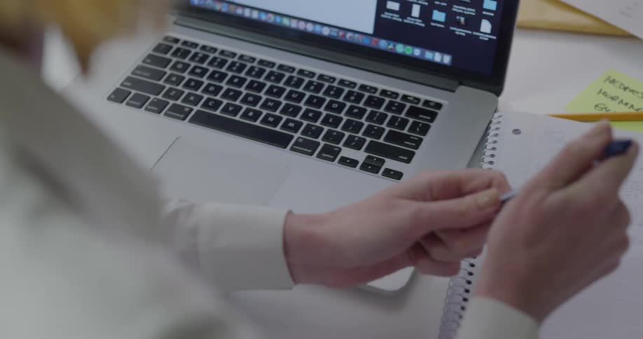 A woman in an office works on her laptop and takes notes