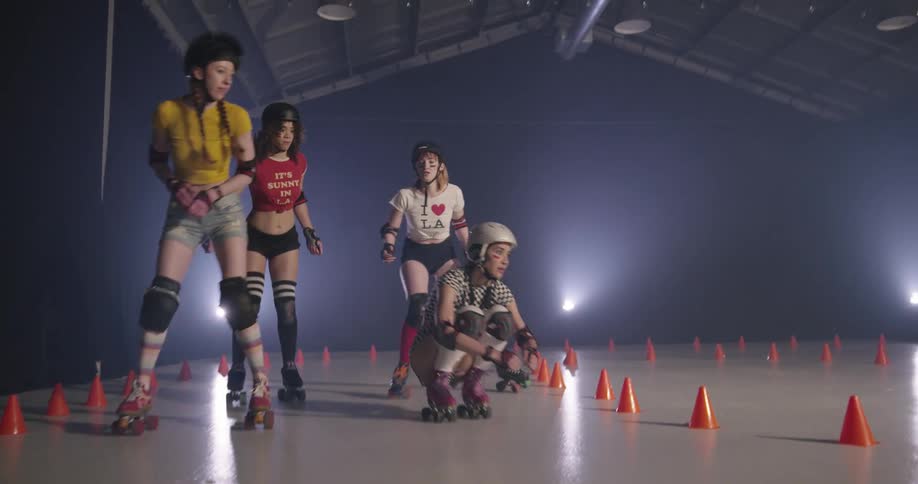 Four women in shorts helmets and pads rollerskate around a small rink with a track marked off with orange cones