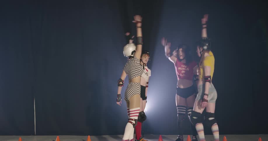 Four women wearing roller skates and protective gear stand in a circle and cheer