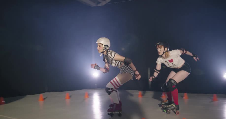 Four women in helmets and pads roller skate around a small rink with a track marked by orange cones