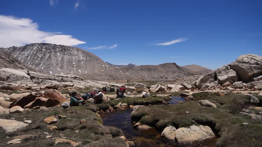 Hikers sitting by a stream of water in the mountains on a sunny day with a few clouds in the sky