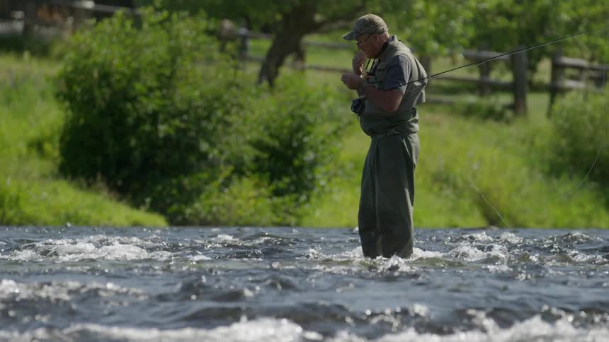 A man in fishing gear stands in a river preparing a lure or hook