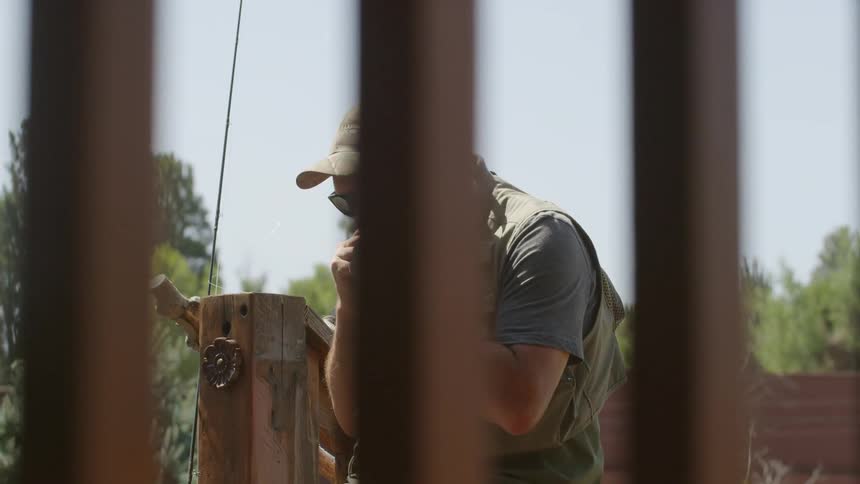 A man in fishing gear sits on a dock preparing a hook