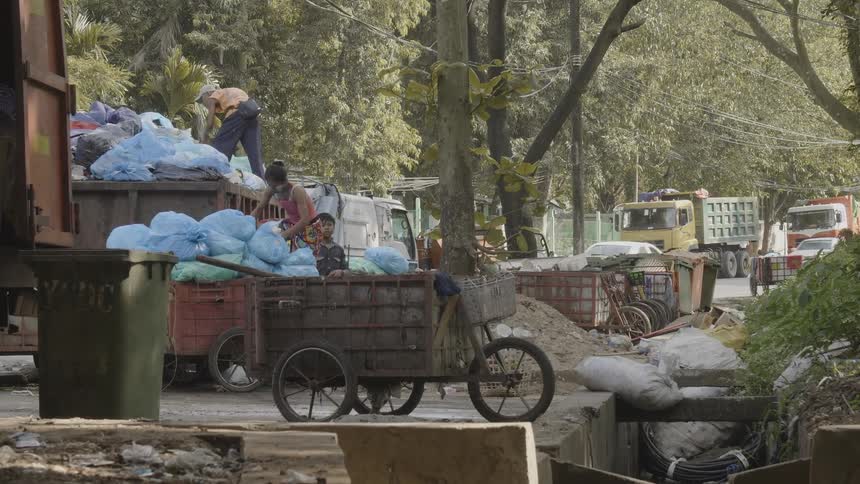 A child stands beside a woman in a truck bed who is throwing blue plastic bags up to a man in a dump truck