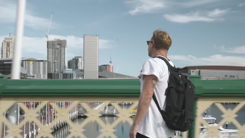 A man wearing a backpack skateboards across a pedestrian bridge over a marina on a bright sunny day